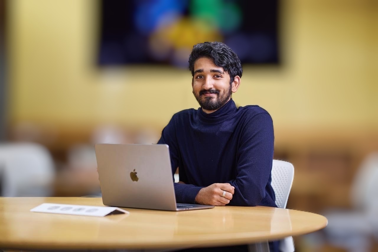 Noah Koshy seated at a table with a laptop, smiling at the camera