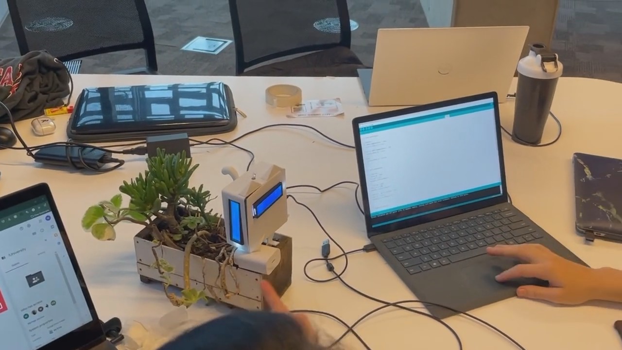 The SproutCat prototype running on a shared table: its planter and glowing LCD sit beside a laptop open on the Arduino code, among other laptops, cables and a water bottle
