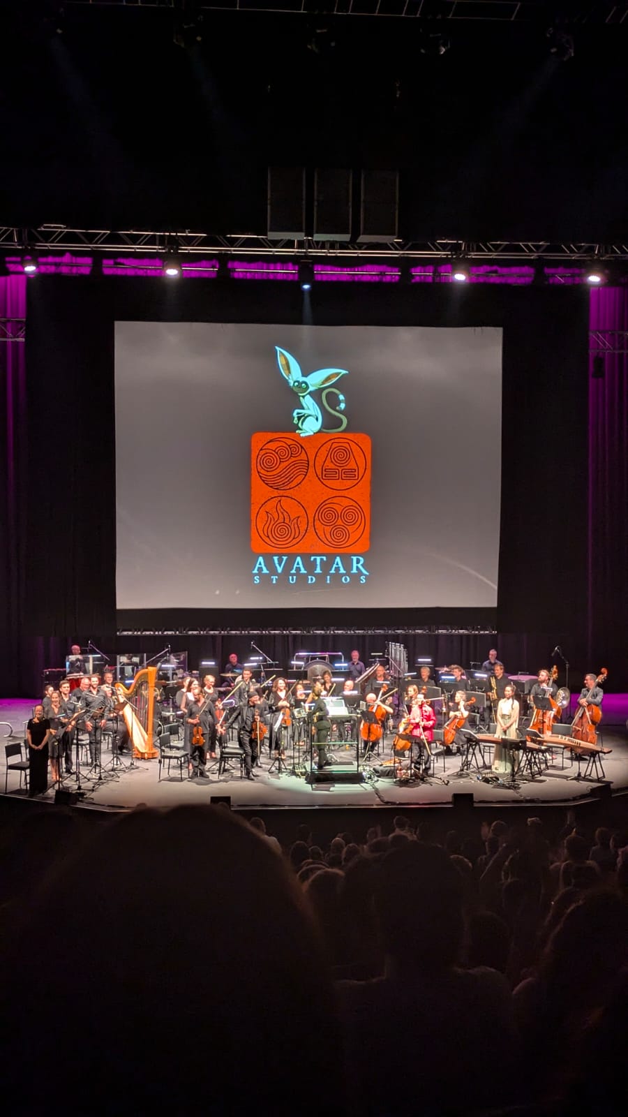 Orchestra on stage beneath a screen showing the Avatar Studios logo, seen from the audience