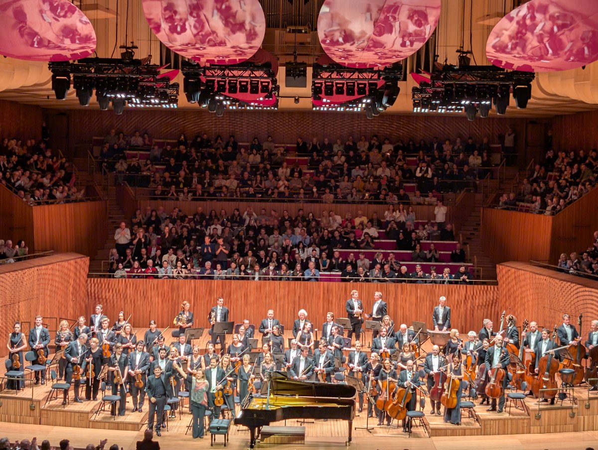Symphony orchestra standing on stage in a wood-panelled concert hall at a Beethoven concert, the audience seated behind them