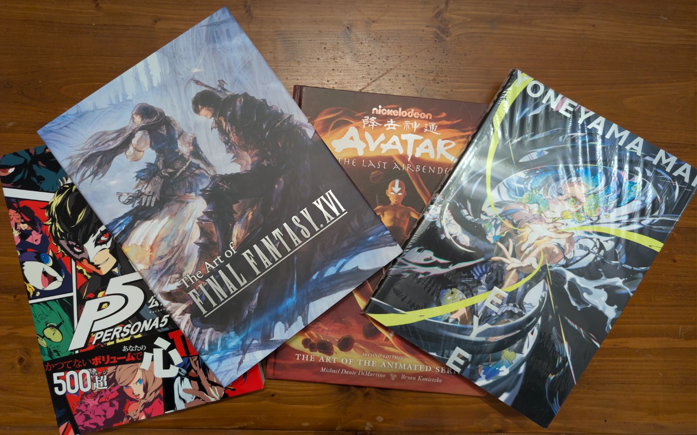 Four art books fanned out on a wooden table: Persona 5, The Art of Final Fantasy XVI, Avatar: The Last Airbender – The Art of the Animated Series, and Yoneyama Mai’s EYE