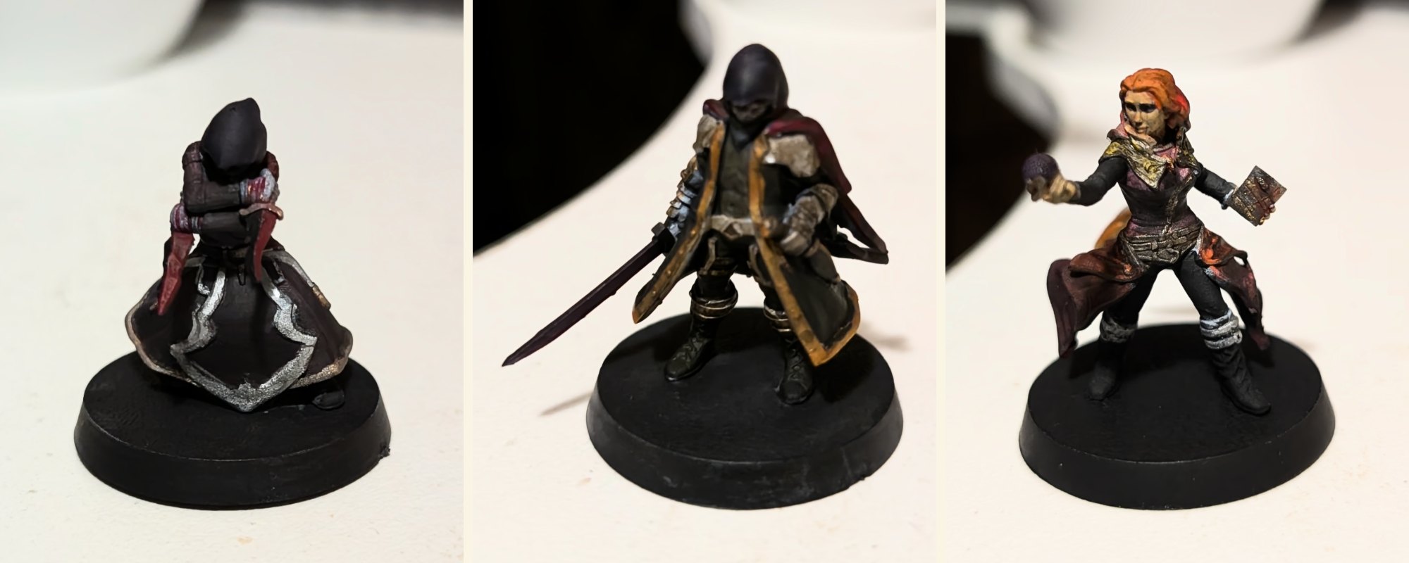 Three hand-painted miniatures on black bases: a hooded figure in a dark robe with silver trim, a hooded swordsman in a gold-edged coat, and a red-haired figure holding an orb and a book